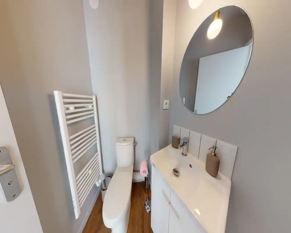 Bagno compatto e moderno con WC, mobile lavabo, specchio rotondo e scaldasalviette. Colori neutri e pavimento effetto legno per un aspetto pulito e minimale.