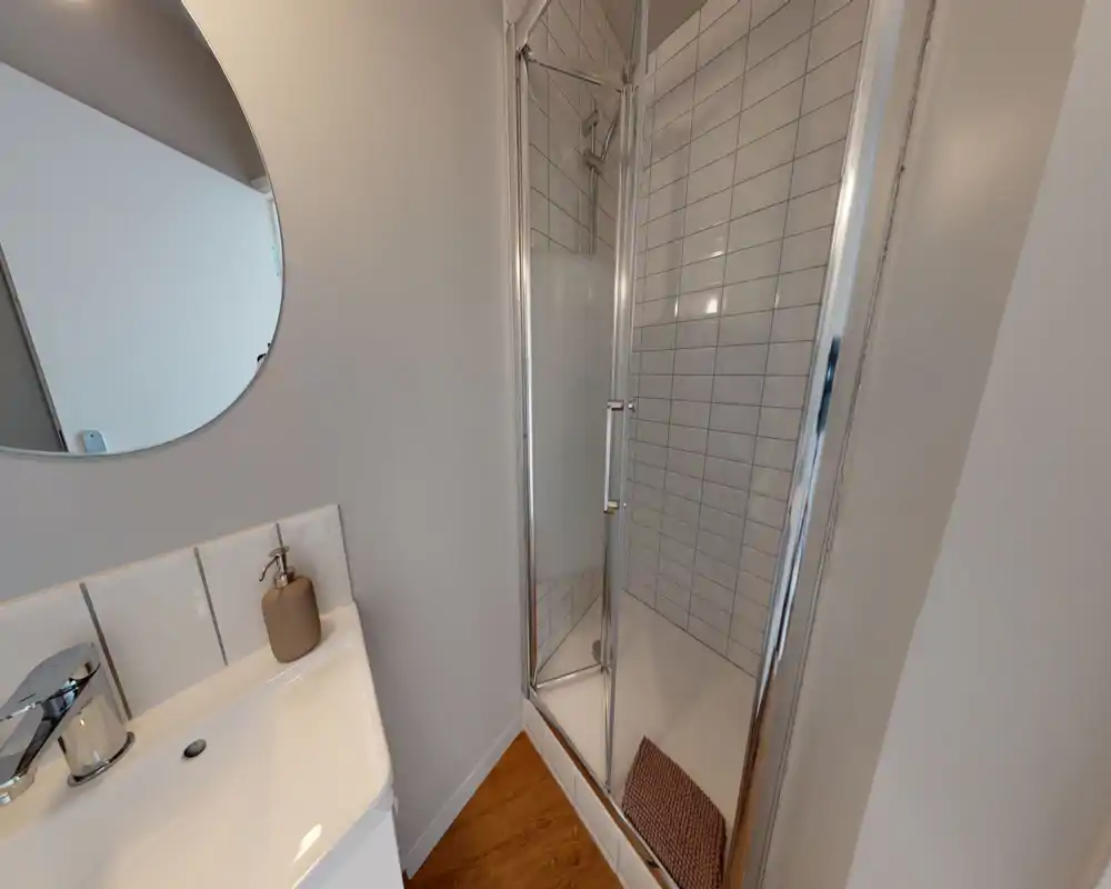 Bagno compatto e moderno con cabina doccia piastrellata, porta in vetro e piccolo lavabo con specchio rotondo: pulito e ben illuminato.