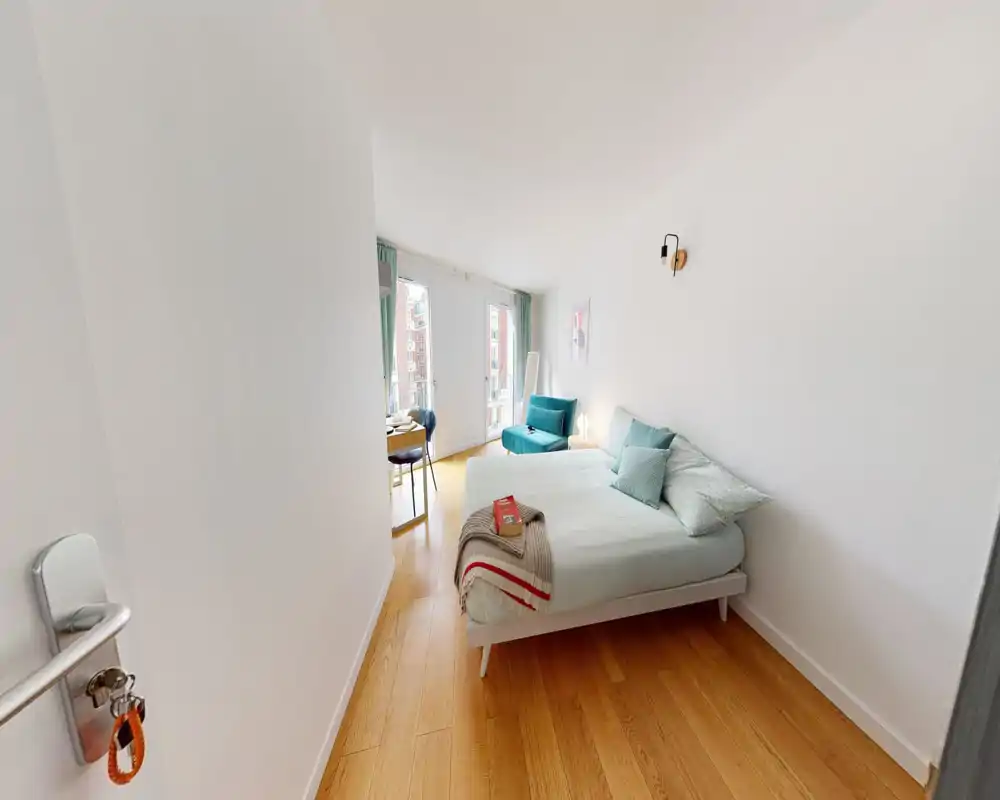 Camera da letto luminosa e moderna con un letto matrimoniale ben rifatto, poltrona color turchese e grandi finestre che lasciano entrare molta luce naturale — perfetta per soggiorni tranquilli.