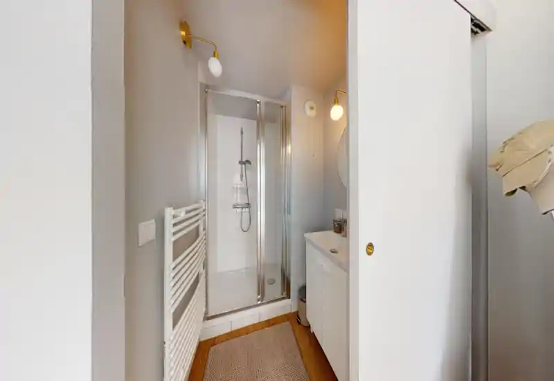 Salle de bains moderne et compacte avec cabine de douche en verre, radiateur porte-serviettes mural et petit meuble vasque. Tons neutres et sol effet bois pour une ambiance propre et accueillante.