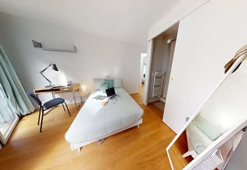 Chambre moderne et lumineuse avec lit double central, petit bureau près de la fenêtre et parquet chaleureux — parfaite pour un séjour confortable ou le télétravail.