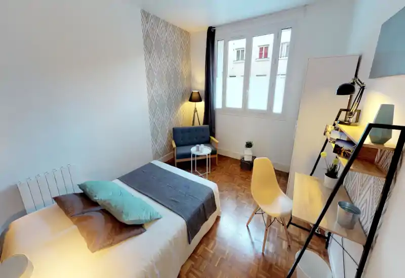Chambre lumineuse et soignée avec un lit double bien fait, coussins décoratifs, petit coin salon et bureau avec étagères — idéale pour un séjour confortable.