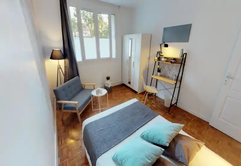 Chambre moderne et lumineuse avec lit bien fait, textiles doux et petit coin salon. La grande fenêtre et des lampes chaudes apportent une atmosphère cosy.