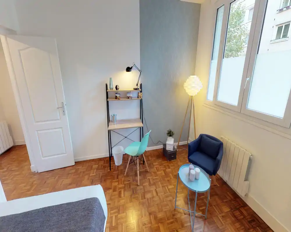 Angolo camera luminoso e ben allestito con il letto in primo piano, una piccola scrivania e una zona conversazione accogliente vicino a una grande finestra — perfetto per annunci che puntano su comfort e luce naturale.