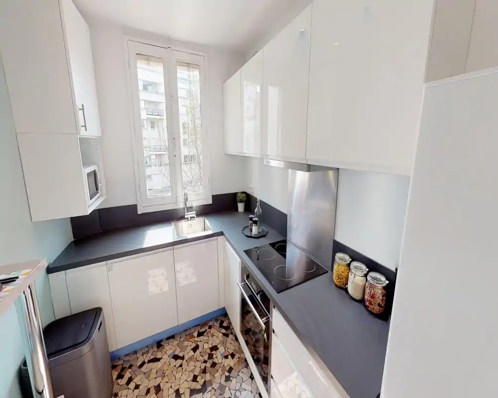 Modern, compact kitchen with glossy white cabinetry, black countertops, induction hob and a window providing natural light. Decorative jars and a small plant add a tidy, welcoming touch.