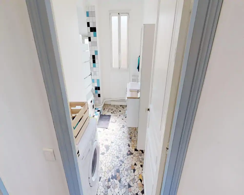 Small bright bathroom with patterned mosaic floor, tiled shower area and a washing machine; white fixtures and natural light from a tall window.