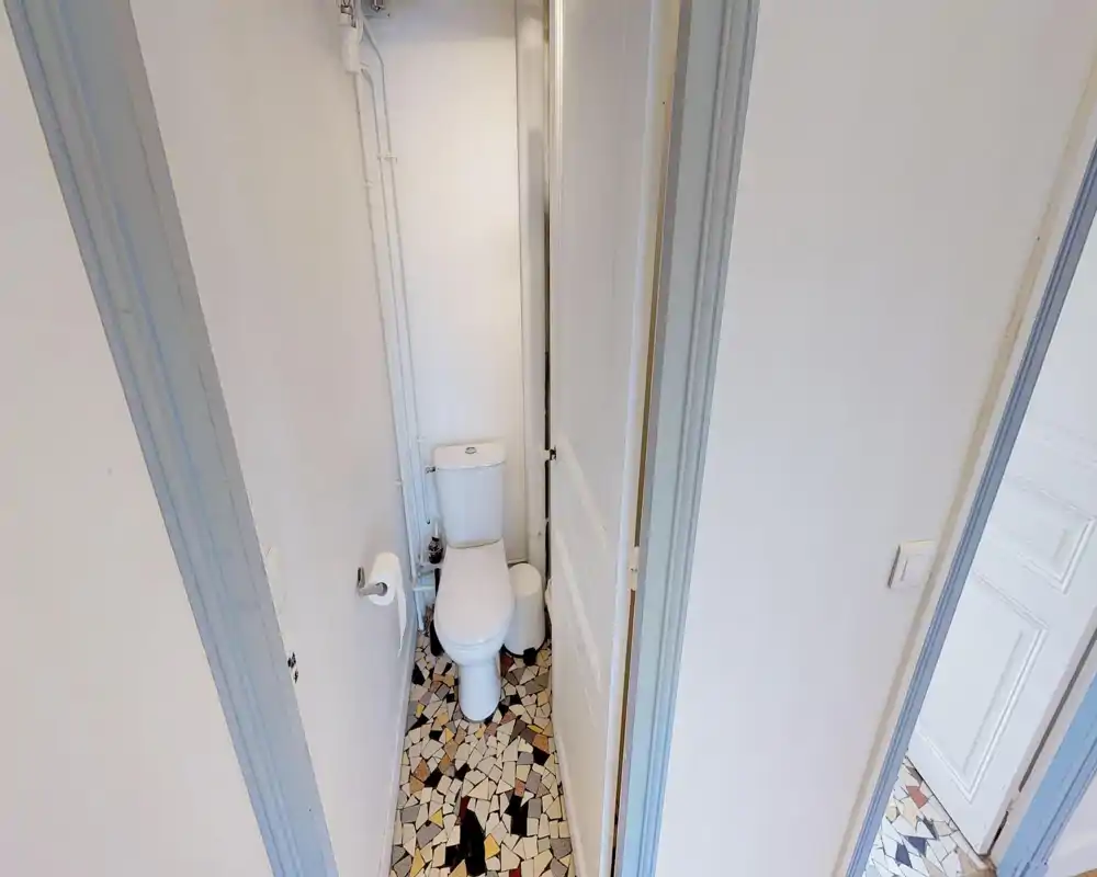 Compact toilet room with a standard white WC, visible plumbing and a patterned mosaic floor. Simple, clean layout suitable for a small apartment.