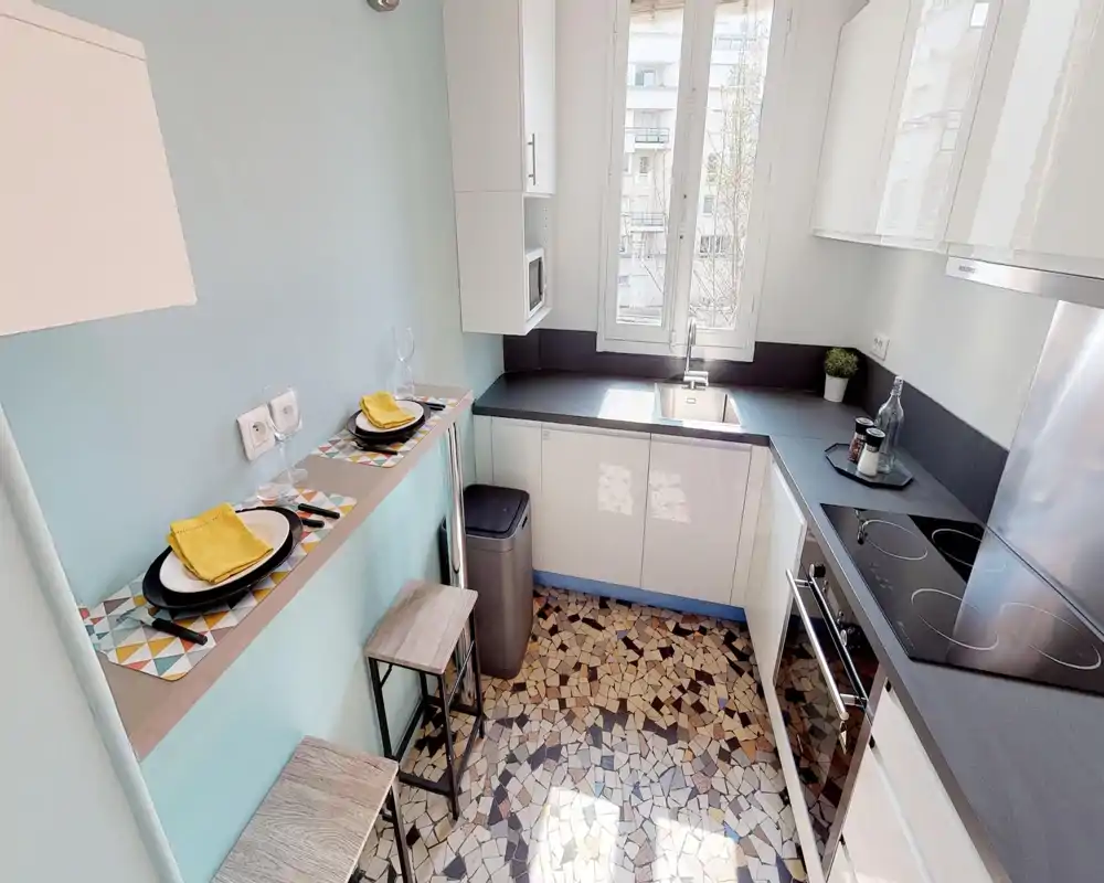 Compact modern kitchen with white cabinets, black countertops and a breakfast bar with stools. Natural light from the window brightens the space and patterned tile flooring adds character.