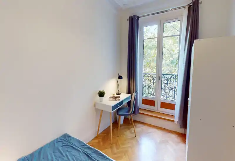 Piccola camera da letto con letto parzialmente visibile, scrivania compatta e sedia vicino a una finestra alta con ringhiera decorativa. Luce naturale abbondante, pavimento in parquet e arredi moderni e minimali, adatta a una persona o a uno studente.
