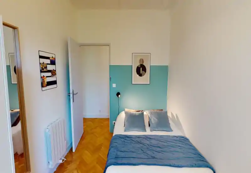 Camera da letto compatta e luminosa con letto matrimoniale con biancheria blu, due cuscini e pannello decorativo nella testata. Pavimento in parquet, lampada da parete e quadretto minimal: perfetta per un soggiorno accogliente.
