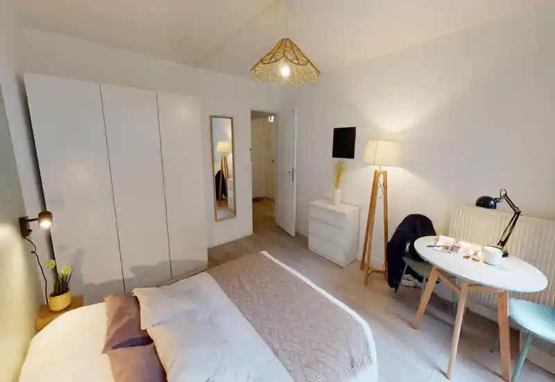 Bright, neatly styled bedroom featuring a large bed with pillows and a textured throw, a wardrobe and a small dining/work table. Warm lamps and neutral tones create a welcoming atmosphere ideal for short-term rentals.