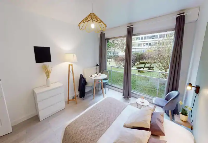 Bright, modern bedroom with a large window overlooking a grassy courtyard. The bed is made with layered cushions and a patterned throw; a small dining table and comfortable seating create a versatile living space.