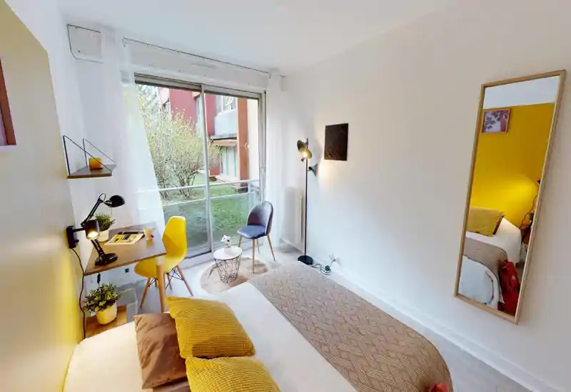 Bright, cozy bedroom with a large window and tasteful yellow accents. The bed is neatly made with textured throws and cushions; a small desk and seating area sit beside the window overlooking greenery.