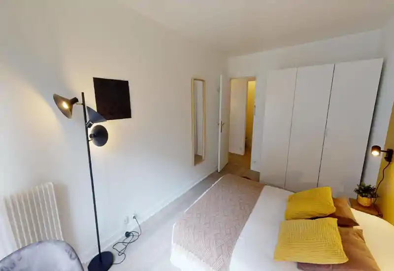Bright, modern bedroom with a double bed dressed in textured linens and yellow accent cushions, a tall wardrobe, full-length mirror and stylish floor lamp.
