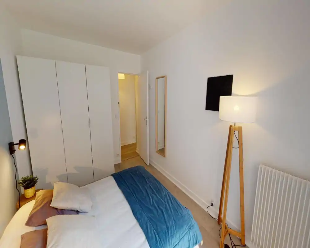 Camera da letto luminosa e ben arredata con un letto matrimoniale in lenzuola neutre e un copriletto blu; armadio, specchio a figura intera e lampada da terra creano un ambiente accogliente.