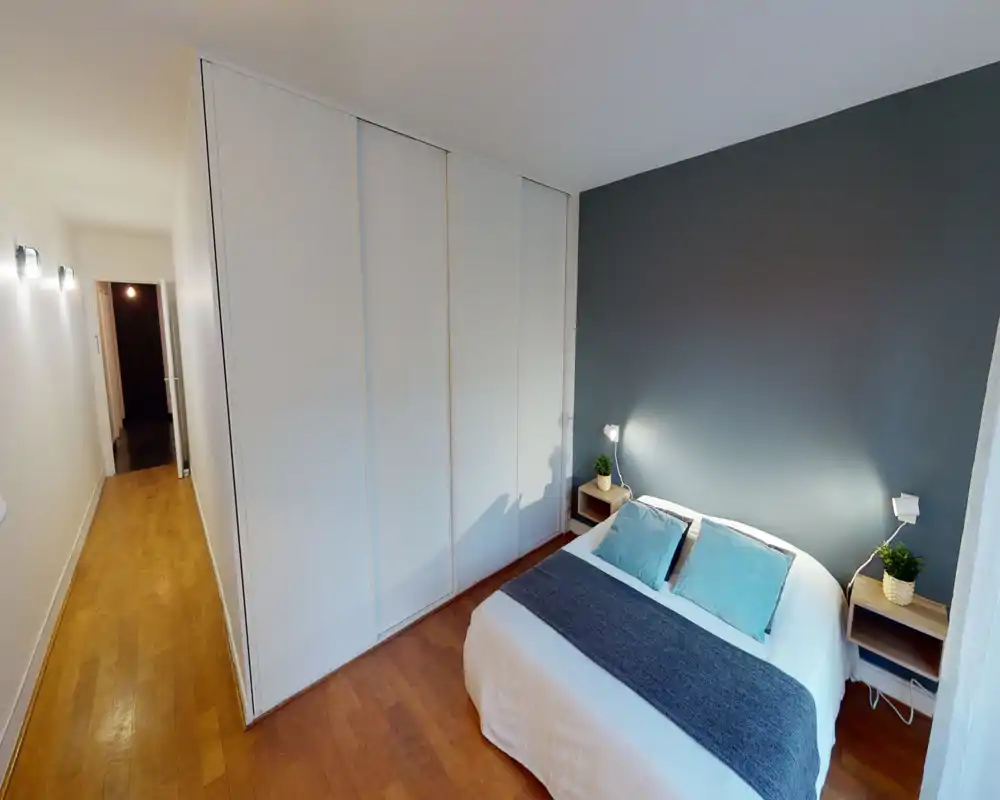 Chambre double soignée avec lit bien fait, tables de chevet équipées de lampes et plantes, et grand placard intégré. Palette neutre et sol en bois chaleureux pour une ambiance moderne et apaisante, idéale pour les annonces.