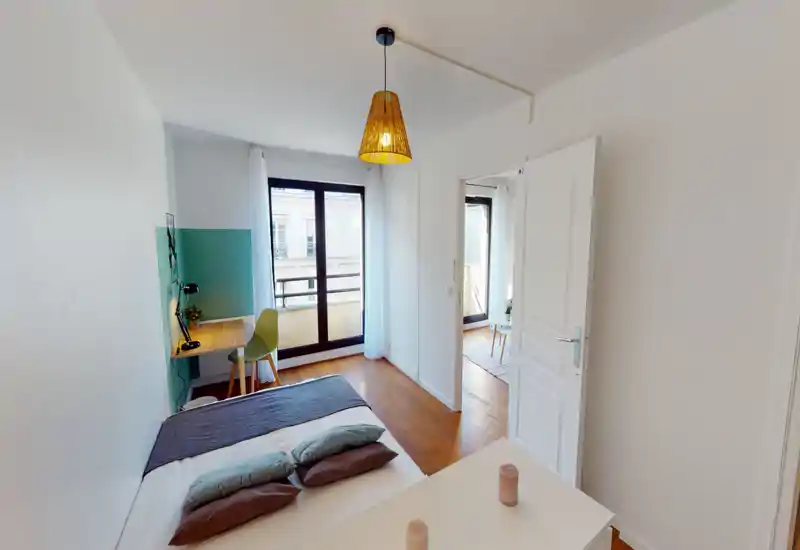 Chambre moderne et lumineuse avec un lit double soigné, coussins coordonnés et un petit bureau près de la fenêtre. Grandes portes-fenêtres donnant sur un balcon apportant une belle luminosité.