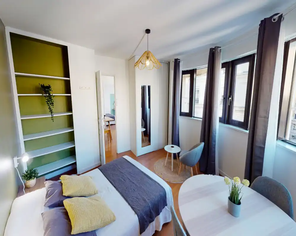Chambre lumineuse et tendance avec un grand lit habillé de linge neutre et coussins, un coin salon cosy près de trois grandes fenêtres et une armoire avec miroir. Lumière naturelle, sol en bois et étagères vertes apportent chaleur.