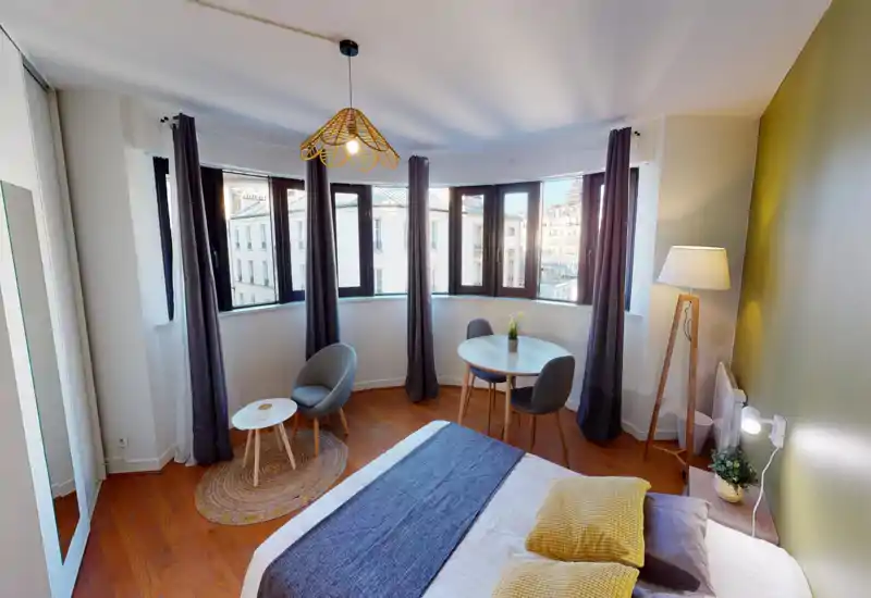 Bright, stylish bedroom with a large bay-window curve, double bed dressed in neutral linens and yellow accent pillows, small seating area and a round dining table — ideal for city stays with lots of natural light.