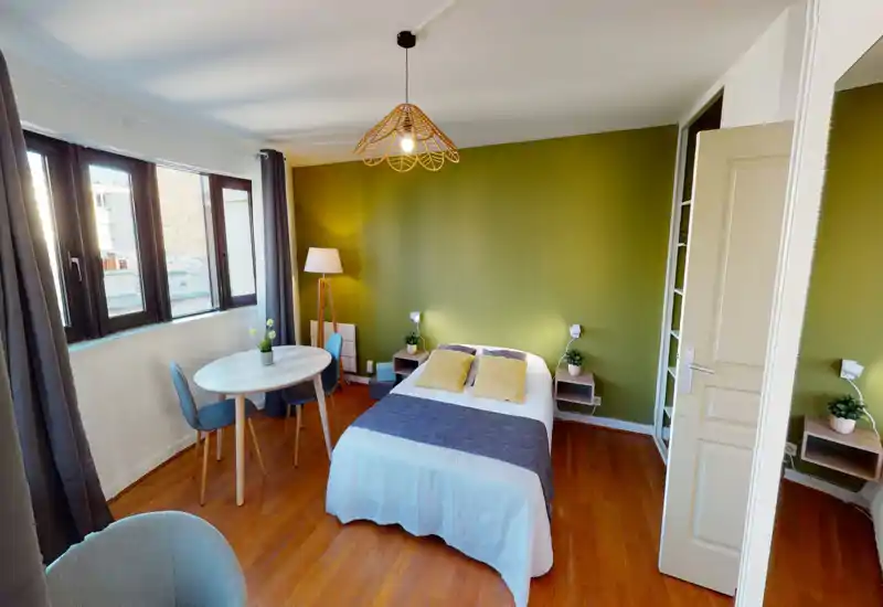 Sunny, stylish bedroom with a comfortable double bed, soft textiles and an olive-green accent wall. Large windows provide abundant natural light and a small dining table and chairs make the space functional for both sleeping and working.