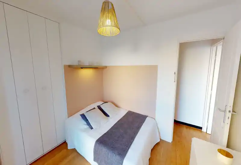 Chambre lumineuse et chaleureuse avec un lit double bien fait, coussins décoratifs et une lampe murale sur étagère. Placard intégré et sol en bois apportent rangement et convivialité.