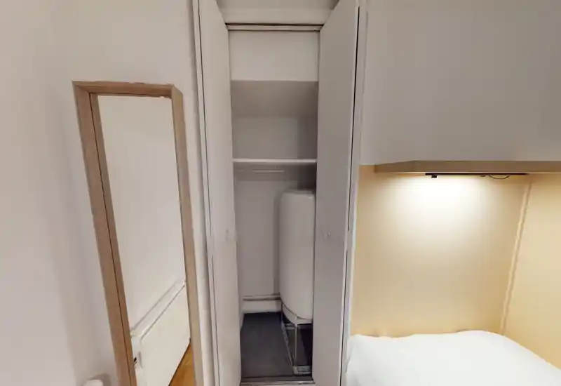 Coin de chambre compact avec une armoire encastrée contenant un chauffe-eau. On aperçoit une partie du lit et une lampe de chevet chaude sur des murs neutres — utile pour signaler un placard technique dans la zone nuit.