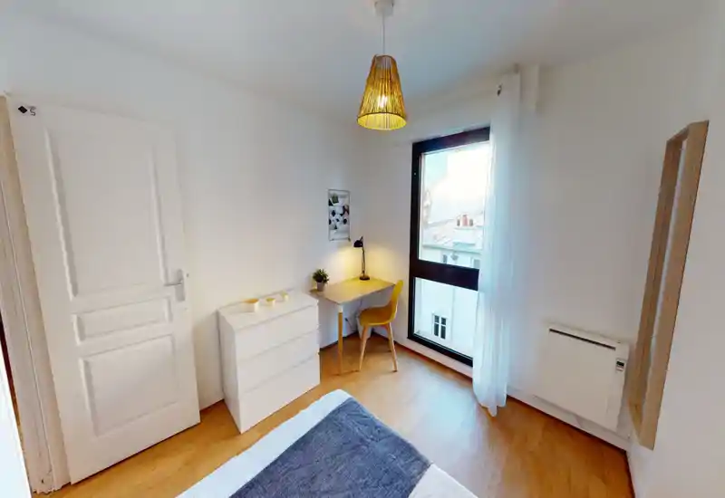 Coin chambre moderne et lumineux avec bord du lit visible, sol en bois, commode compacte et petit bureau près d'une grande fenêtre — lumineux et bien mis en scène pour une annonce.