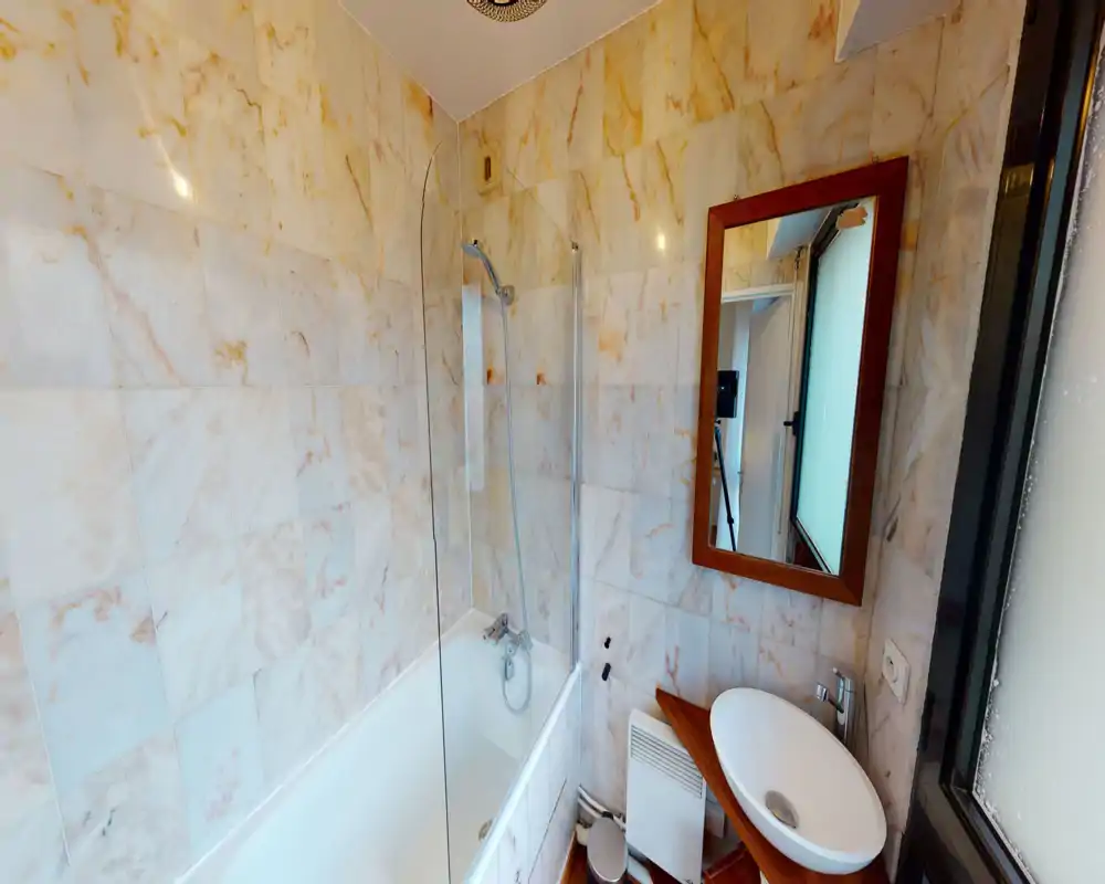 Salle de bains lumineuse entièrement carrelée avec baignoire et paroi de douche, lavabo rond sur plan et miroir à cadre en bois. L'espace est bien éclairé et net sur la photo.