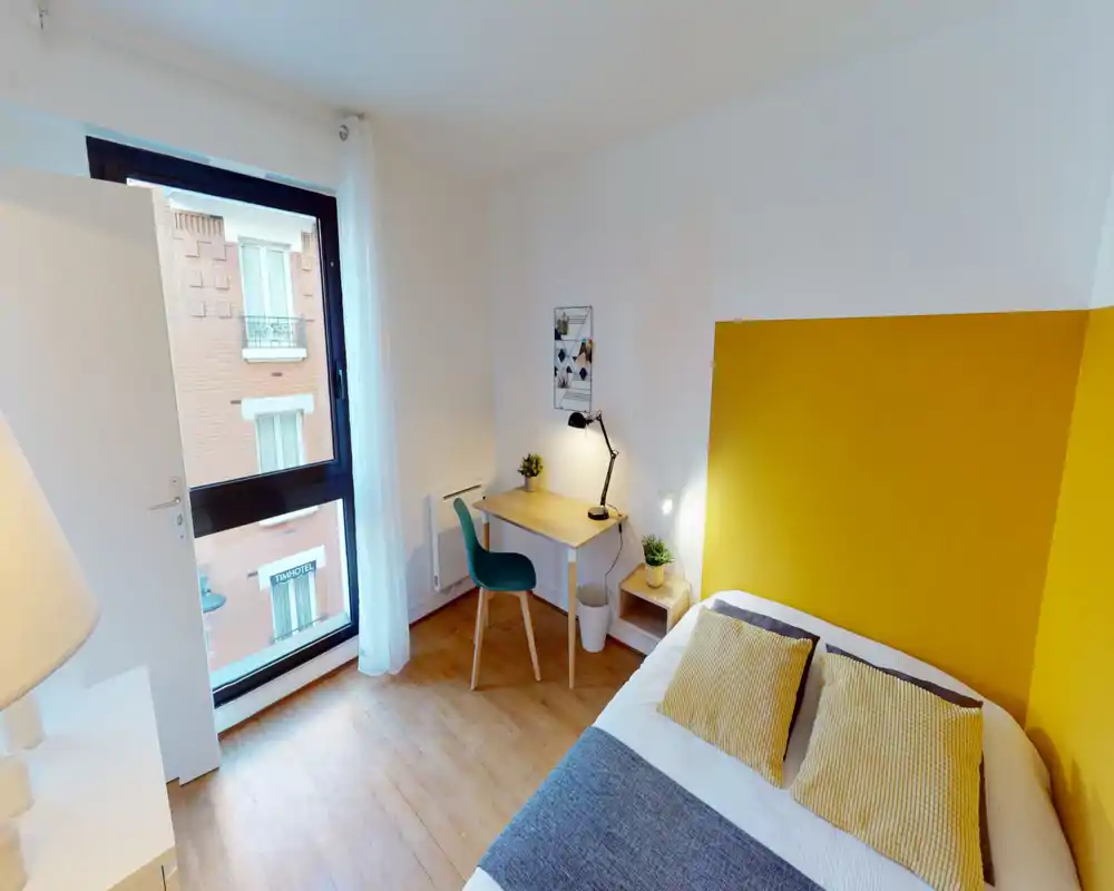 Chambre moderne et lumineuse avec un mur jaune, lit double avec coussins assortis et un petit bureau près de la fenêtre — parfaite pour courts séjours ou voyageurs d'affaires.