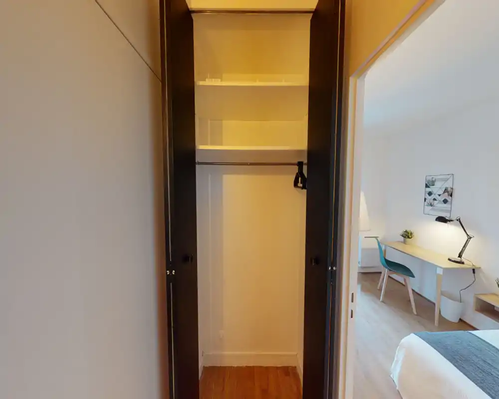 Vue d'une petite chambre montrant un placard ouvert au premier plan et un lit bien fait avec un bureau visible par la porte. Décor minimal, sol en bois et lumière agréable — espace fonctionnel adapté aux séjours courts ou individuels.