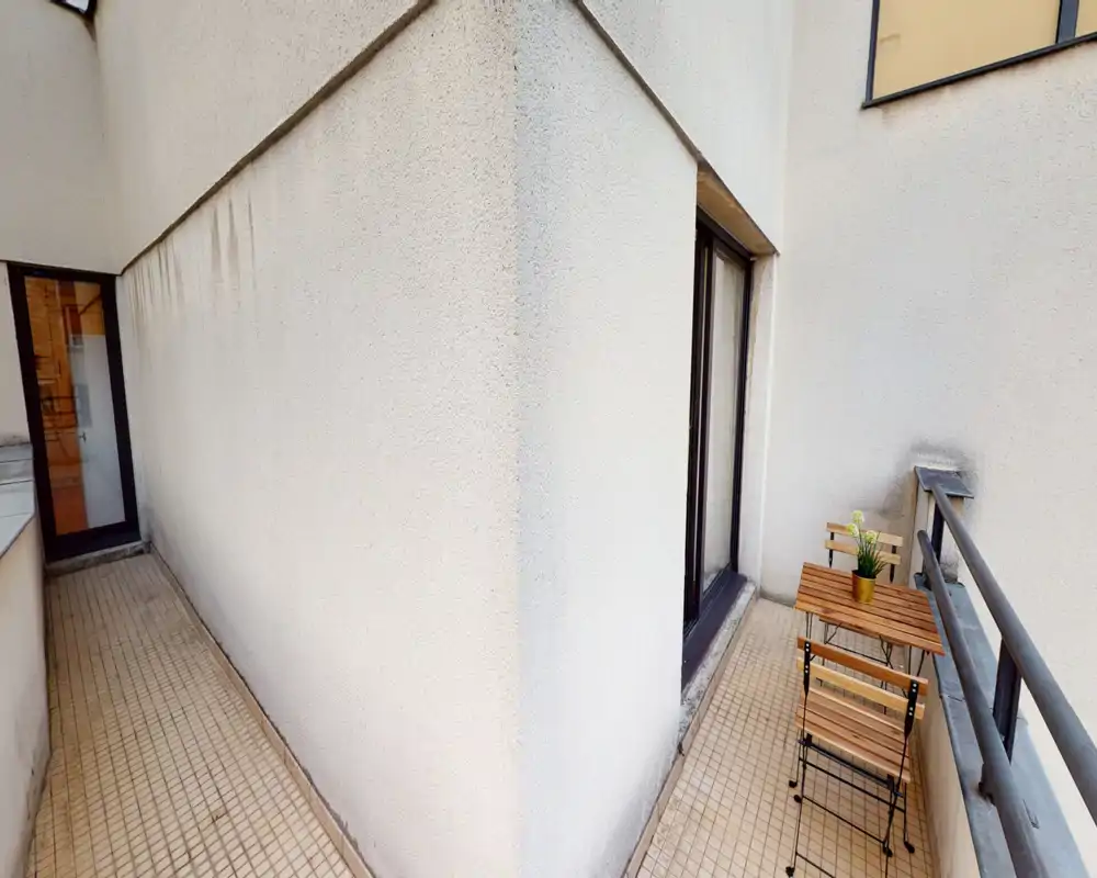 Petit balcon privé étroit avec sol carrelé, rambarde métallique et une petite table et chaises en bois de style bistrot — un coin extérieur compact pour le café du matin.