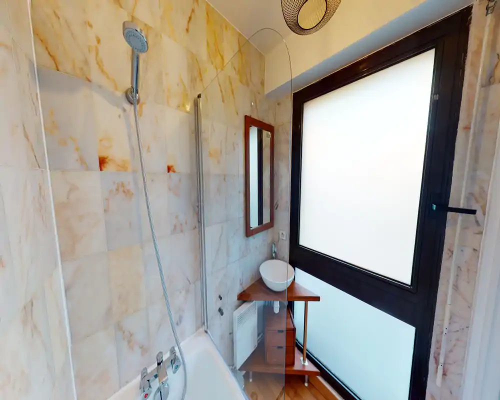 Salle de bains lumineuse carrelée avec baignoire et douchette, meuble d'angle compact avec vasque et une fenêtre dépoli assurant intimité et lumière naturelle.