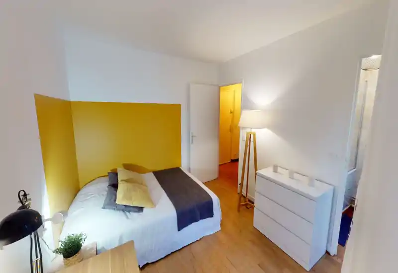 Chambre lumineuse et moderne avec un grand lit, un mur d'accent jaune et des textiles douillets ; bien mise en scène avec lampadaire et commode.