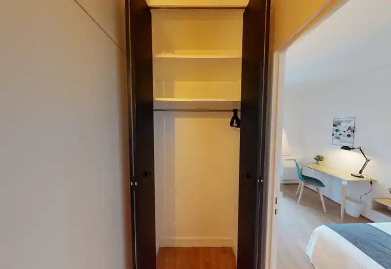 Vue d'une petite chambre montrant un placard ouvert au premier plan et un lit bien fait avec un bureau visible par la porte. Décor minimal, sol en bois et lumière agréable — espace fonctionnel adapté aux séjours courts ou individuels.