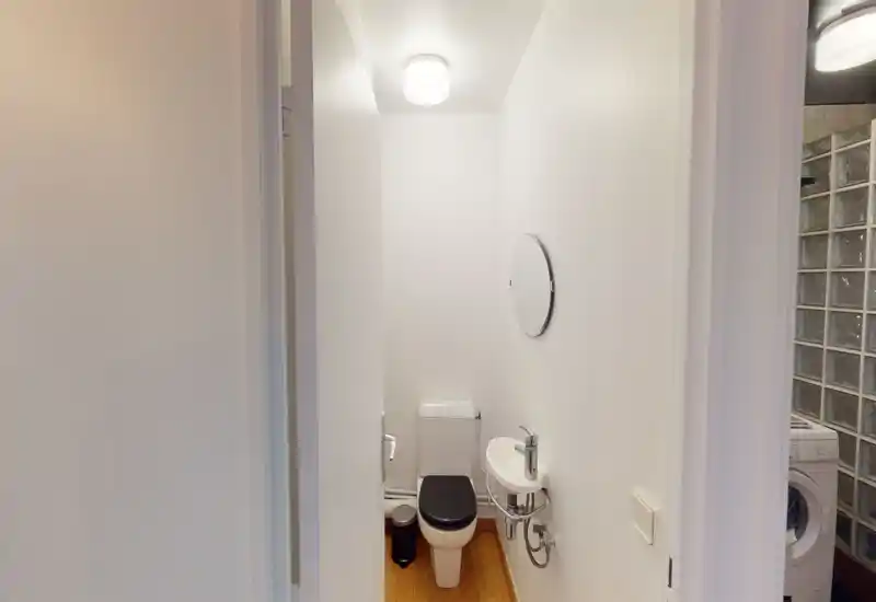 Compact WC with a toilet, small wall-mounted basin and round mirror; bright white walls and wood-effect floor create a clean, minimalist look.