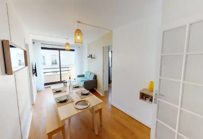Bright, modern living area with a dining table set for four, a compact sofa and large floor-to-ceiling window leading to a balcony; warm wooden floors and woven pendant lights create a cozy atmosphere.