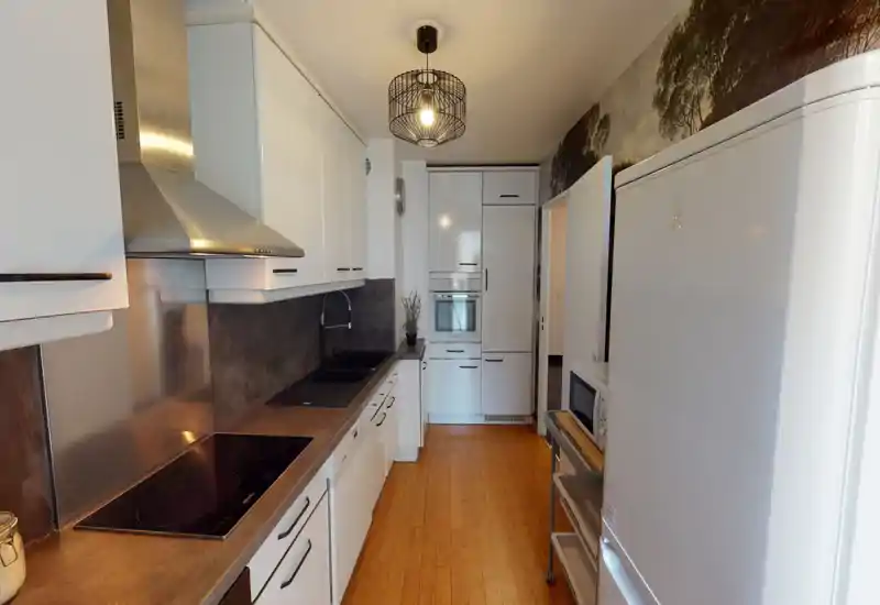 Modern galley kitchen with white cabinets, integrated appliances, induction cooktop and a stainless hood. Warm wood flooring and tasteful lighting create an inviting cooking space.