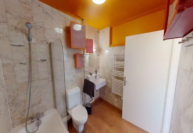 Modern small bathroom with bathtub and showerhead, wall-mounted sink, toilet and heated towel rack; warm yellow ceiling and marble-effect wall tiles give character.