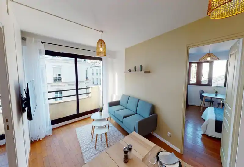 Bright, stylish living room with a compact sofa, nesting coffee tables and large sliding windows that open to a small balcony. The space is well staged with warm lighting and wooden floors, ideal for a cozy city apartment.
