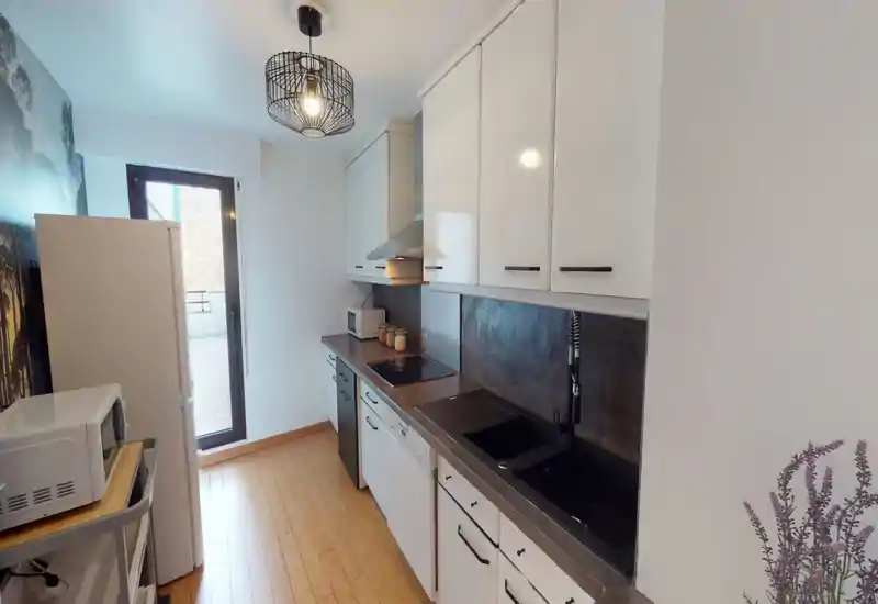 Bright modern galley kitchen with white cabinetry, dark countertop and integrated appliances; wood flooring and a glass door to a small balcony provide natural light.