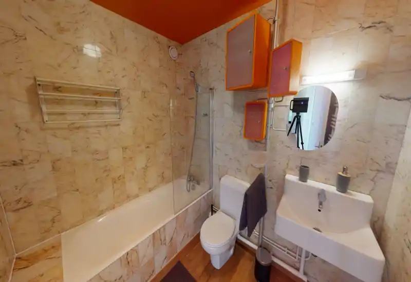 Modern marble-tiled bathroom with bathtub and shower screen, wall-mounted sink, toilet and mirrored cabinet; warm orange accents and good lighting make the space inviting.