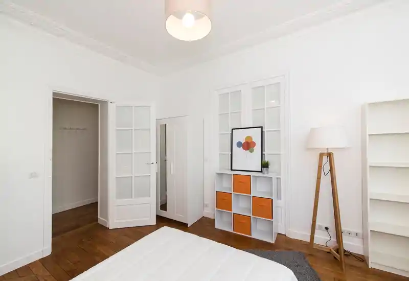 Chambre moderne et lumineuse avec un lit blanc bien fait au premier plan, parquet, une armoire blanche et un meuble à cubes aux accents orange accompagné d'une lampe sur trépied.