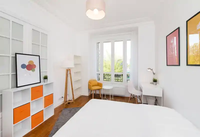 Chambre moderne et lumineuse avec grande fenêtre, linge de lit blanc et touches de couleur chaleureuses. La pièce comprend un bureau, des étagères et un fauteuil jaune confortable, idéale pour une location en ville.