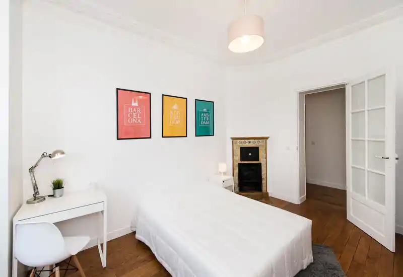 Chambre lumineuse et minimaliste avec un lit double centré, un petit bureau blanc et une chaise, des affiches décoratives et une cheminée carrelée ancienne — propre, aérée et bien éclairée.