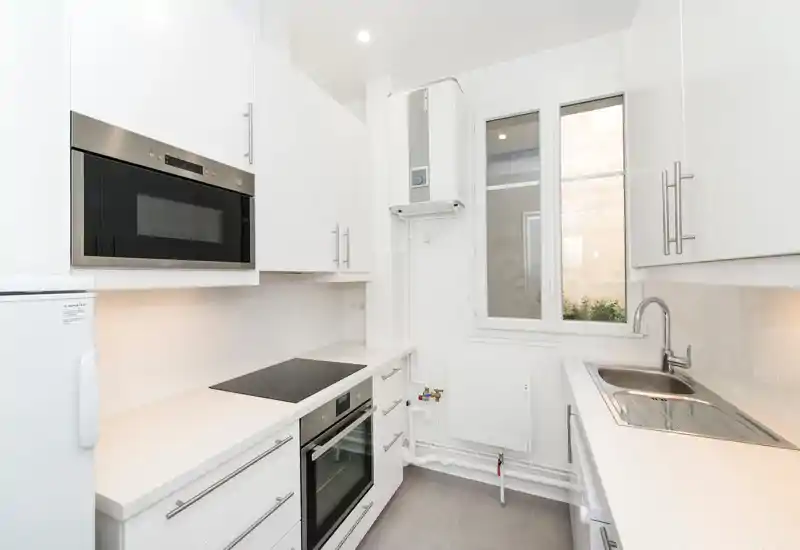 Cuisine moderne et lumineuse avec placards blancs, électroménager inox, plaque à induction et four encastré; design épuré et minimal, évier près de la fenêtre.