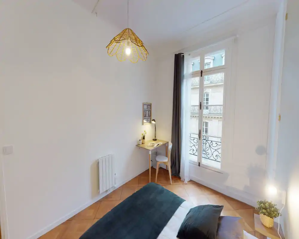 Chambre lumineuse et minimaliste avec lit au premier plan, petit bureau près d’une haute fenêtre et éclairage soigné pour une ambiance calme et aérée.