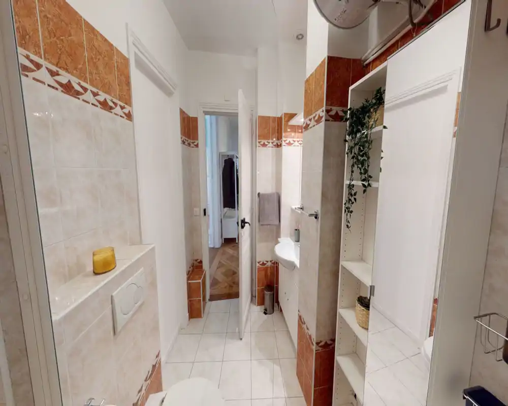 Salle de bains carrelée avec lavabo blanc, bouton de chasse mural et étagères. Espace propre et bien éclairé avec une frise décorative et un agencement compact menant au couloir.