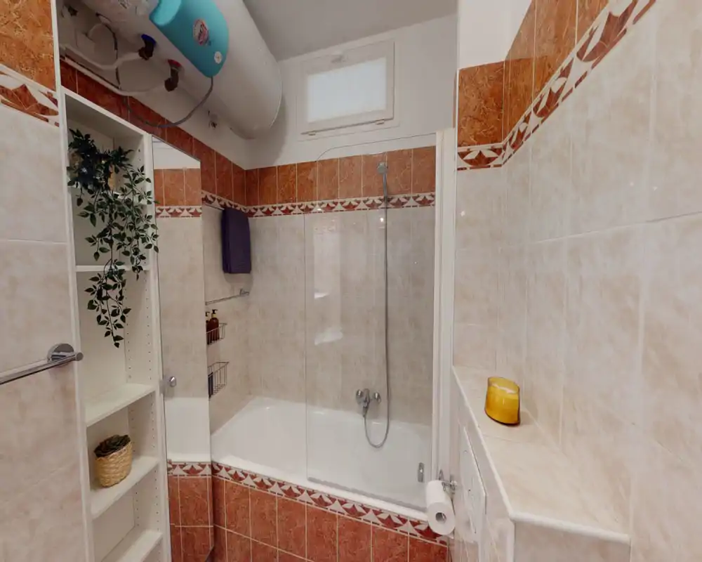 Salle de bains lumineuse et propre avec baignoire-douche, carrelage mural et une étagère fine contenant plantes et articles de toilette.