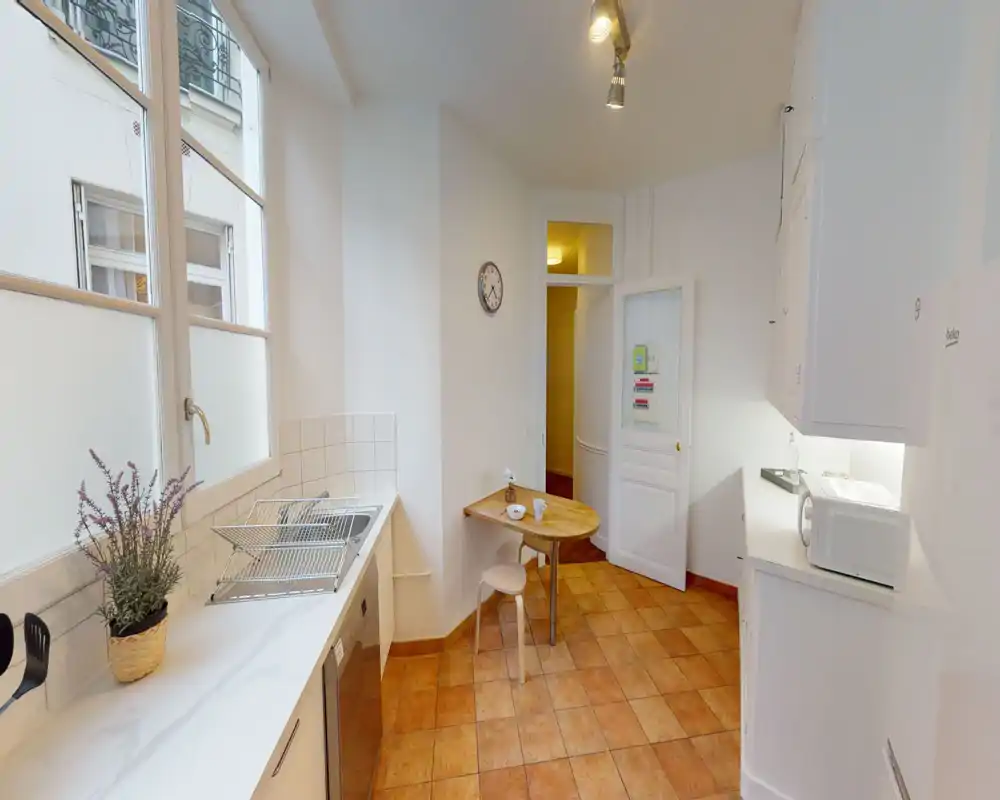 Cuisine compacte et lumineuse avec placards blancs, évier sous une grande fenêtre, micro-ondes sur le plan de travail et petite table en bois — parfaite pour une personne ou un couple.
