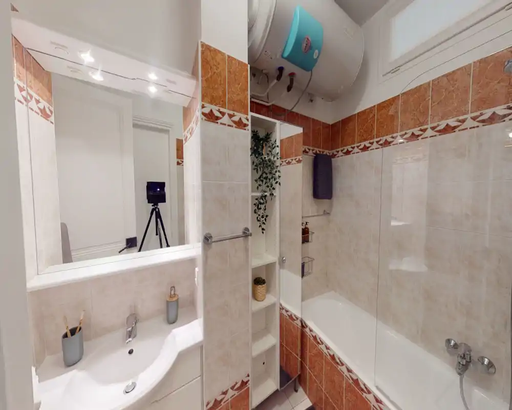 Salle de bains propre et bien éclairée avec lavabo, miroir, étagères intégrées et baignoire avec paroi en verre. Carrelage beige et terracotta pour une ambiance chaleureuse et soignée.